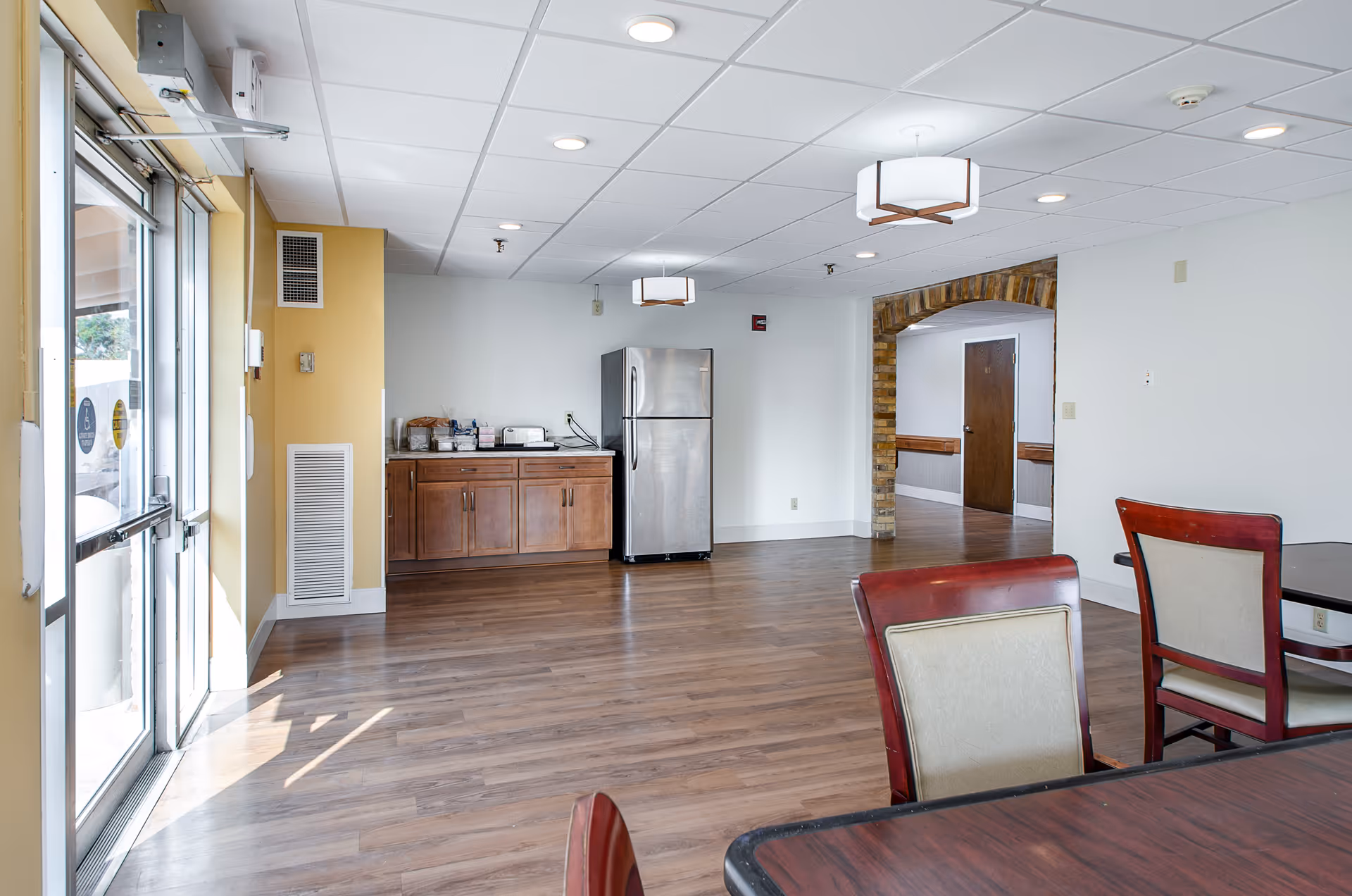 Interior view of a senior living facility room with wooden flooring, a stainless steel refrigerator, wooden cabinets, and tables with chairs. The room has white walls, a yellow accent wall near the entrance door, and ceiling lights. There is an arched doorway with exposed brick leading to another hallway.