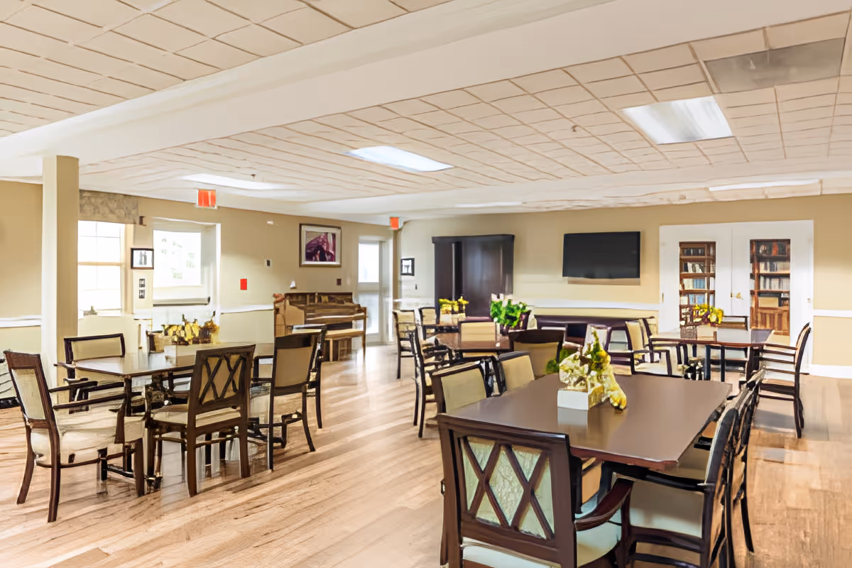 A spacious, well-lit common area with multiple wooden tables and chairs arranged neatly. The room features light-colored walls, a wooden floor, a piano against one wall, a flat-screen TV mounted on another wall, and a bookshelf behind glass doors. There are floral centerpieces on the tables and large windows letting in natural light.