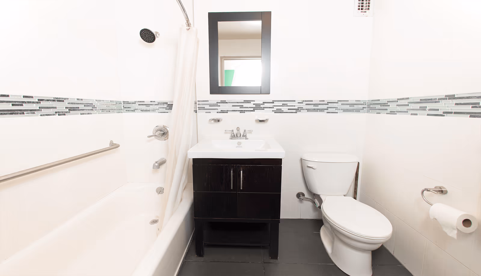 A clean and modern bathroom with a white bathtub and shower combination on the left, a dark wood vanity with a white sink and a rectangular mirror above it in the center, and a white toilet on the right. The walls are white with a decorative horizontal strip of gray and black tiles. There is a silver grab bar on the wall next to the bathtub and a toilet paper holder with a roll of toilet paper on the right wall.