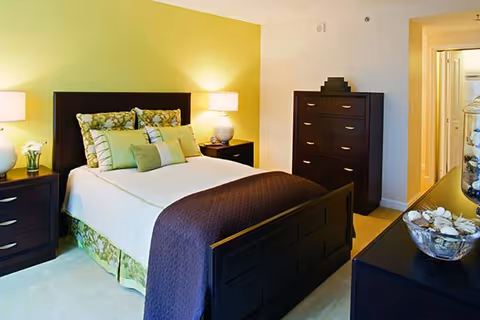 A neatly made bed with a dark wooden frame and headboard, adorned with multiple pillows in green and floral patterns. The room has a yellow accent wall behind the bed, two bedside tables with lamps, a dark wooden dresser, and a hallway leading to another room. The overall decor is warm and inviting.