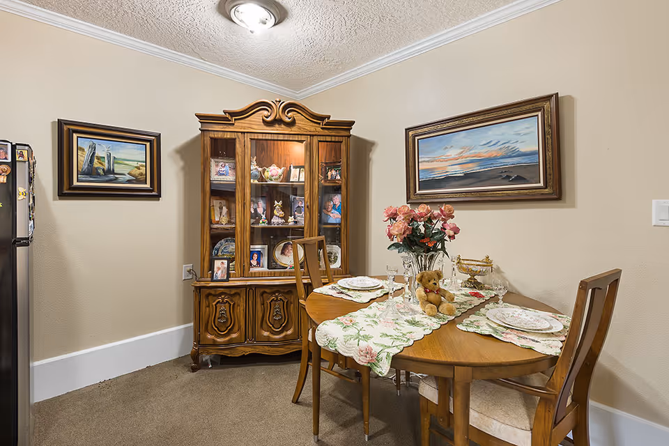 Round wooden dining table set with plates and a floral centerpiece in a cozy room with a wooden china cabinet and framed paintings on the walls.