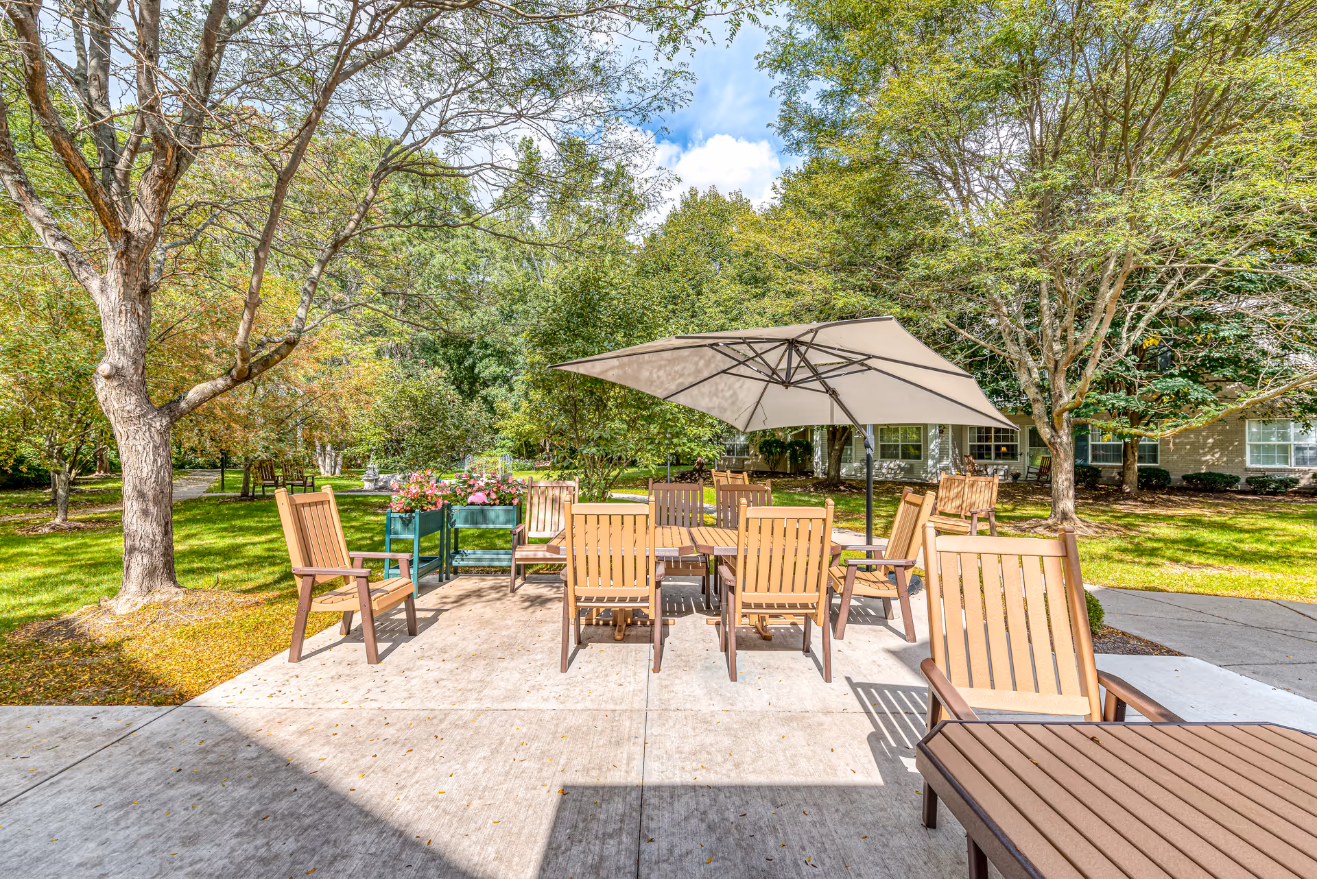 Outdoor patio with wooden chairs and a central table under a large umbrella surrounded by trees and lawn in front of the building.