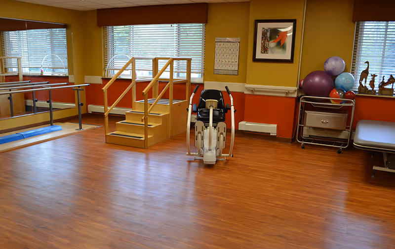 A physical therapy or exercise room with wooden flooring, a set of stairs with handrails, an exercise bike, exercise balls on a metal cart, a treatment table, large windows with blinds, and a wall mirror with ballet bars.