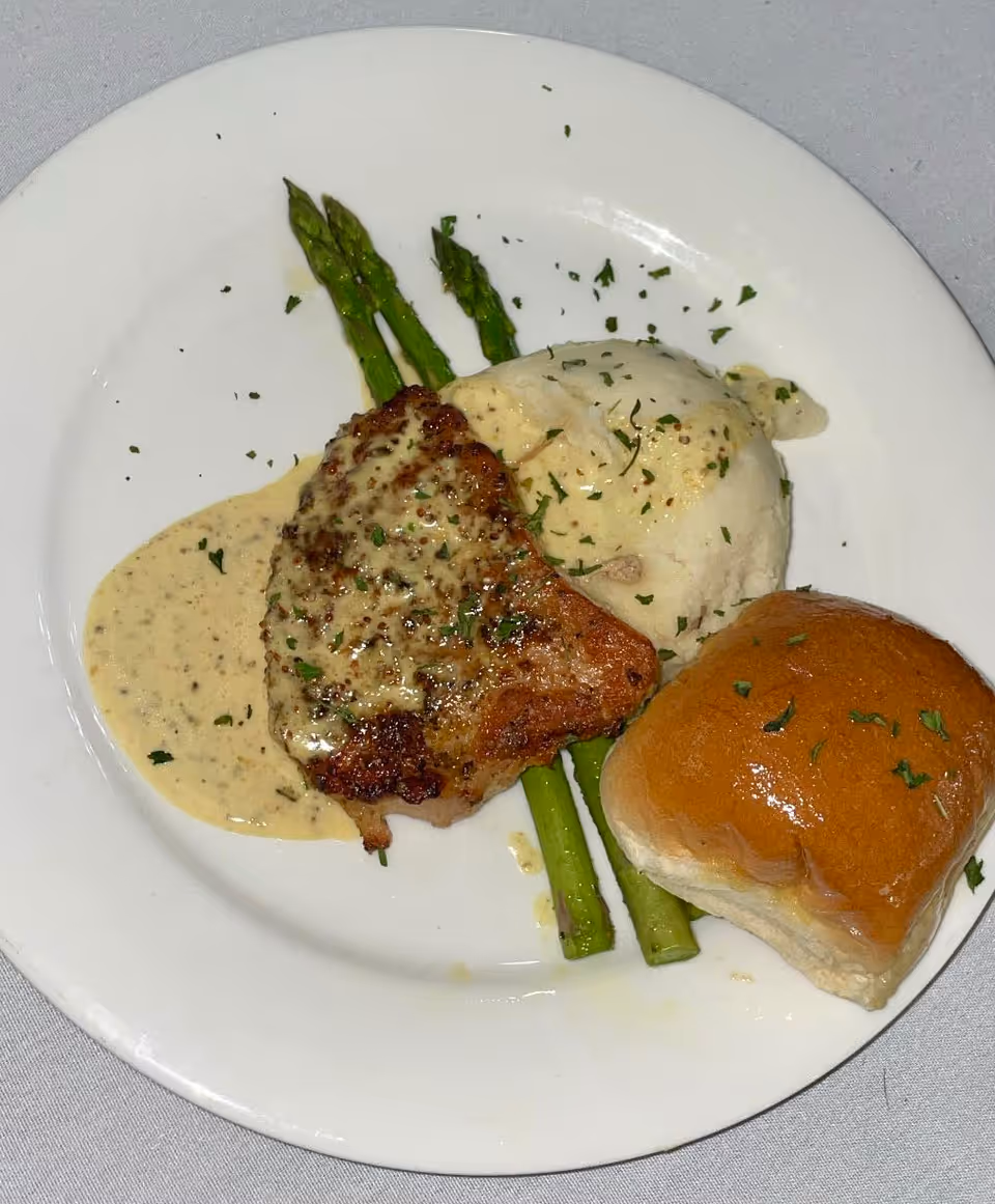 A white plate with a serving of grilled meat topped with creamy sauce, three spears of asparagus, a scoop of mashed potatoes with gravy, and a dinner roll garnished with herbs.