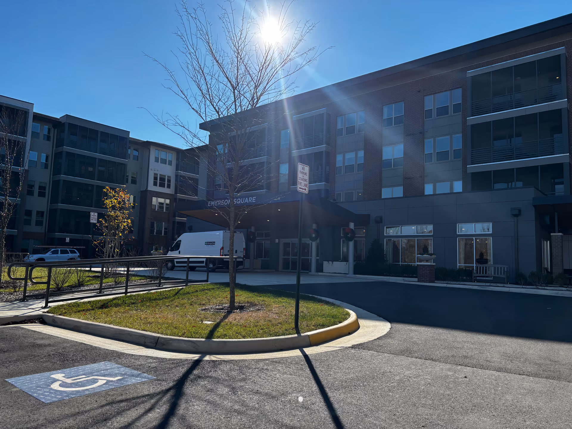 Exterior front entrance of a multi-story senior living building with a driveway, a white van under the entrance canopy, a painted handicap parking symbol in the foreground, and bright sunlight overhead.