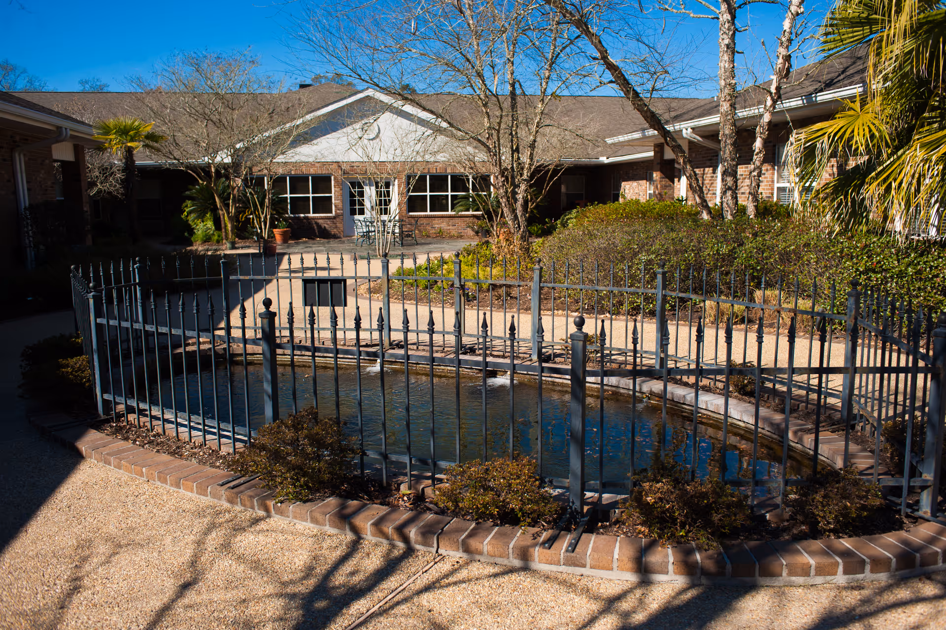 Outdoor courtyard area of a senior living facility with a small fenced pond surrounded by brick edging, shrubs, and trees. The building with large windows and a patio area is visible in the background under a clear blue sky.