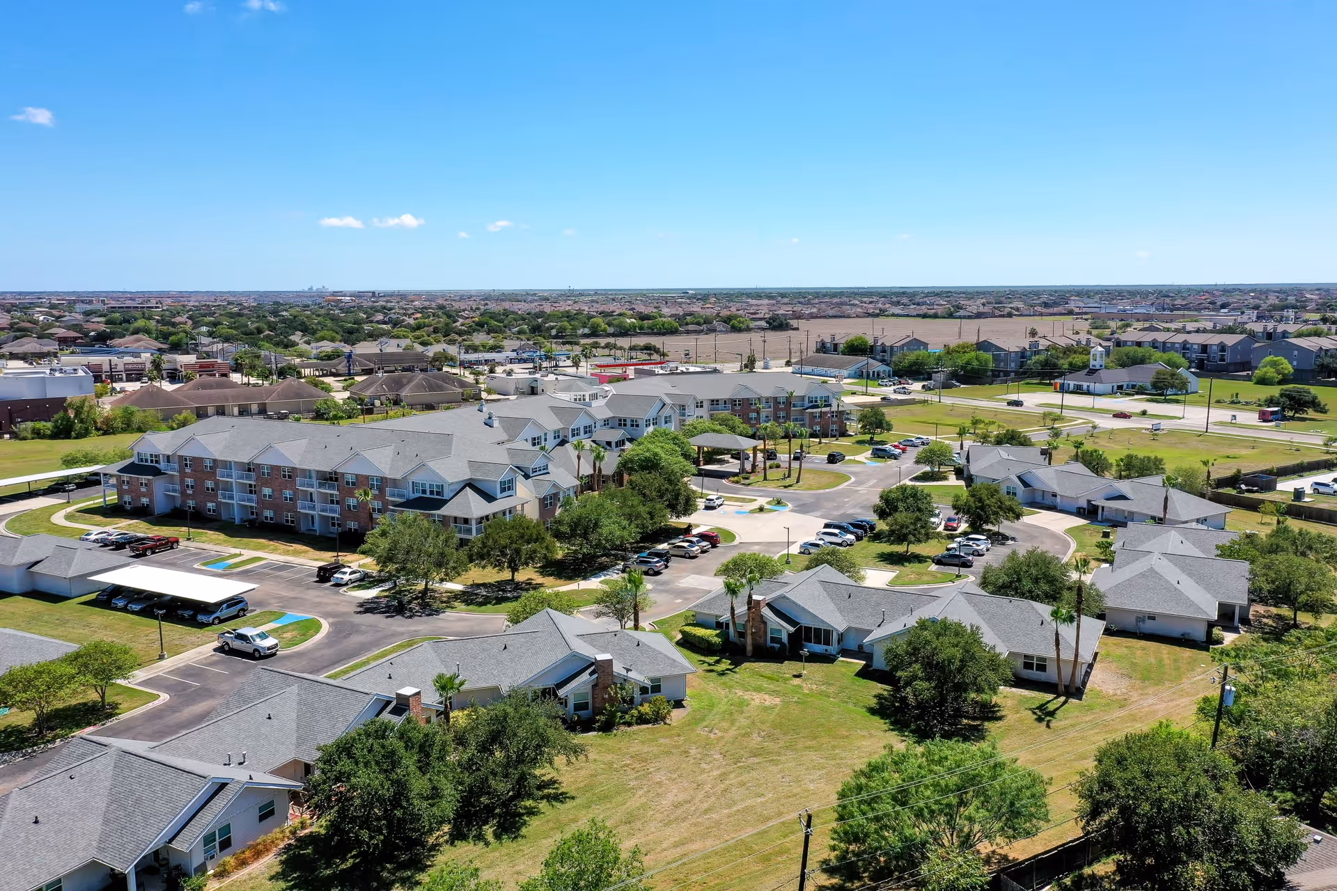 Aerial view of Solstice Senior Living at Corpus Christi showing multiple residential buildings with gray roofs, parking lots with cars, green lawns, trees, and a clear blue sky in the background.
