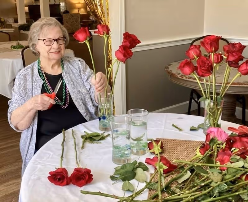 An elderly woman with glasses and a gray cardigan is sitting at a round table arranging red roses in a glass vase. The table is covered with a white tablecloth and has several rose stems, leaves, and three glasses of water on it. The background shows a dining area with tables and chairs.