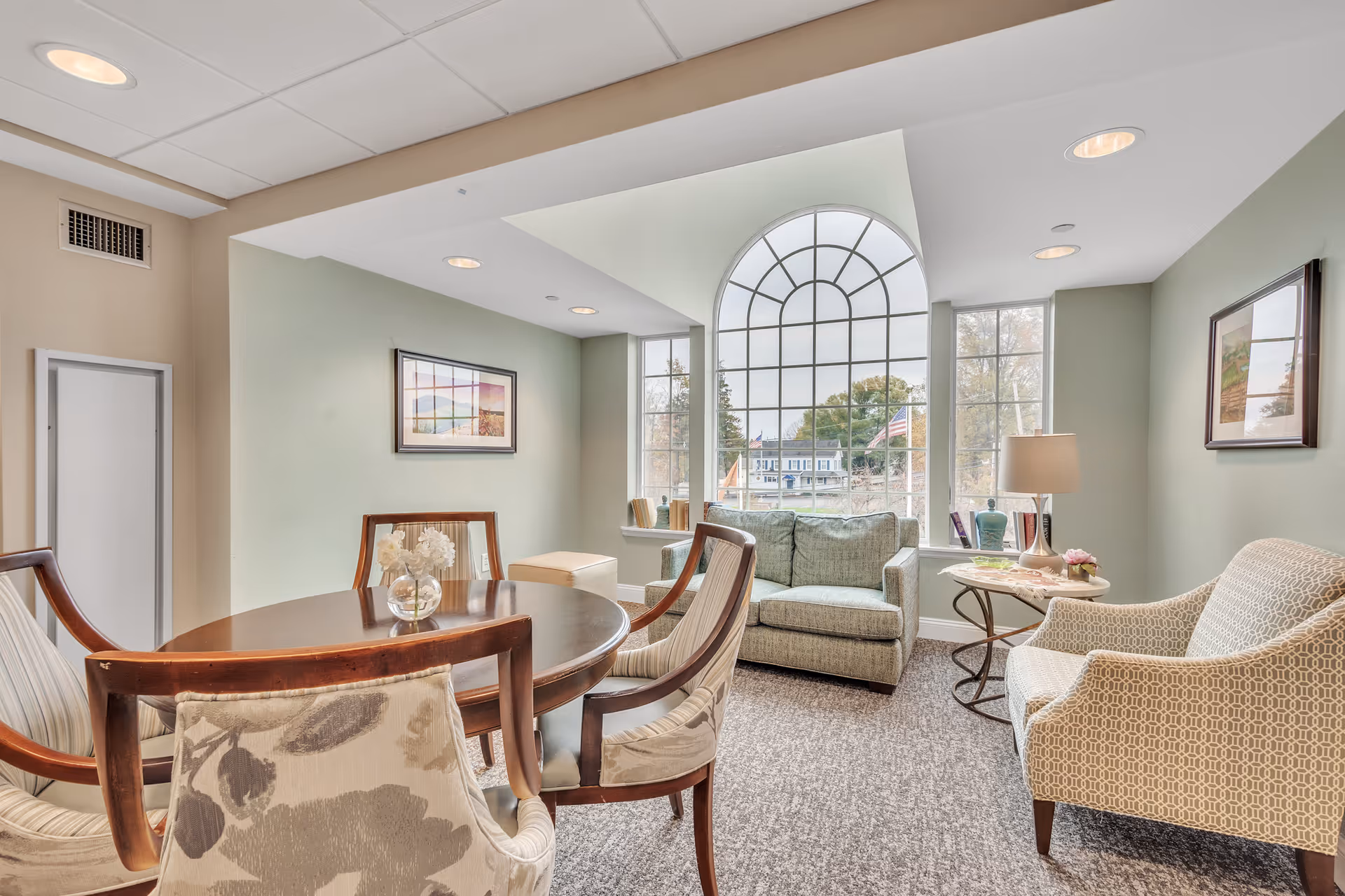 A cozy sitting area in a senior living facility with a round wooden table surrounded by four upholstered chairs, a small loveseat, and an armchair. The room features a large arched window letting in natural light, light green walls, framed artwork, a side table with a lamp and decorative items, and a carpeted floor.