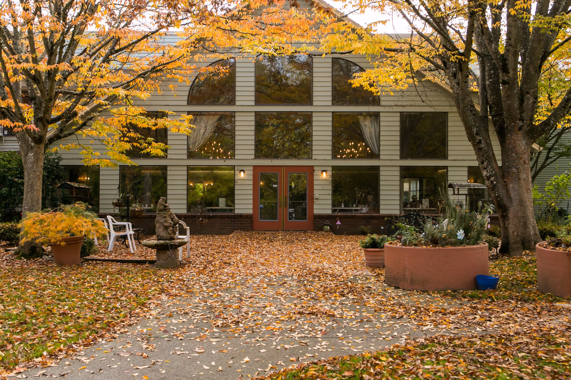 Exterior view of a senior living facility building with large windows and double doors, surrounded by autumn trees with yellow and orange leaves. There are potted plants, a stone fountain, and white chairs on a leaf-covered pathway leading to the entrance.