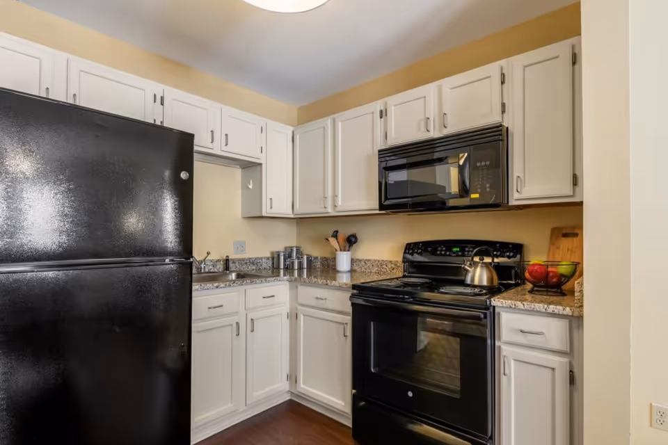 A compact kitchen with white cabinets, granite countertops, a black refrigerator, black electric stove with oven, and a black microwave mounted above the stove. Kitchen utensils are in a white holder on the counter, and a fruit basket with apples and pears is on the right side of the counter.