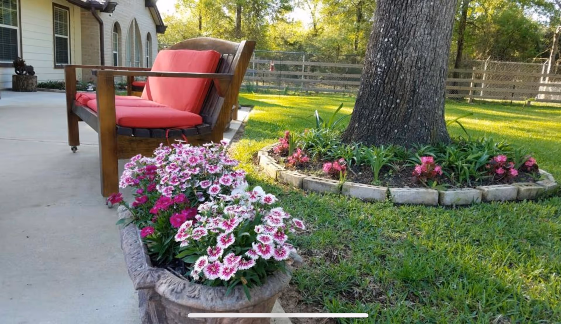 Patio with a wooden bench with red cushions, potted pink-and-white flowers, and a tree surrounded by a flower bed on a grassy lawn.