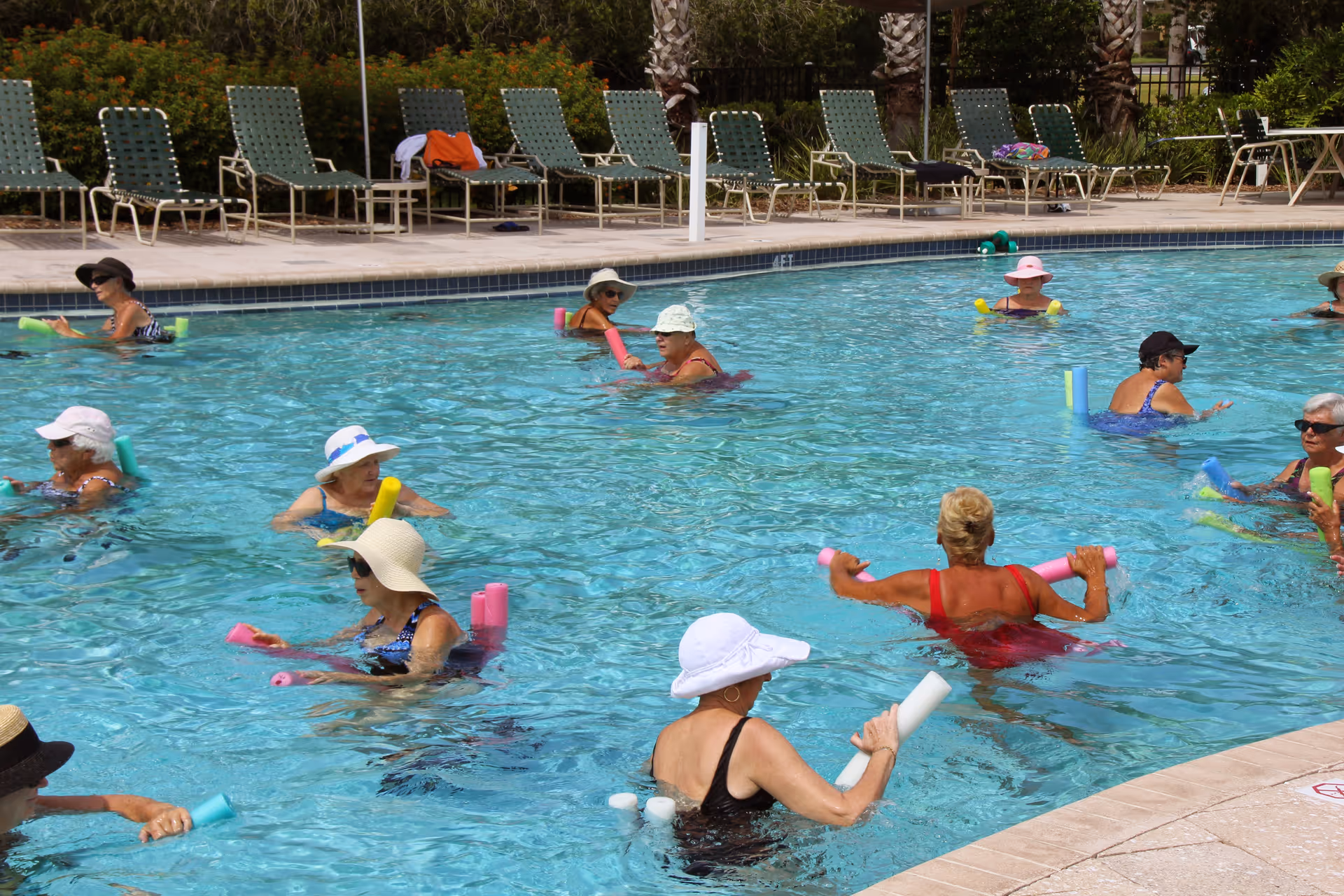 A group of elderly women participating in a water exercise class in an outdoor swimming pool. They are using colorful pool noodles and wearing sun hats and swimsuits. Lounge chairs and greenery are visible around the pool area.