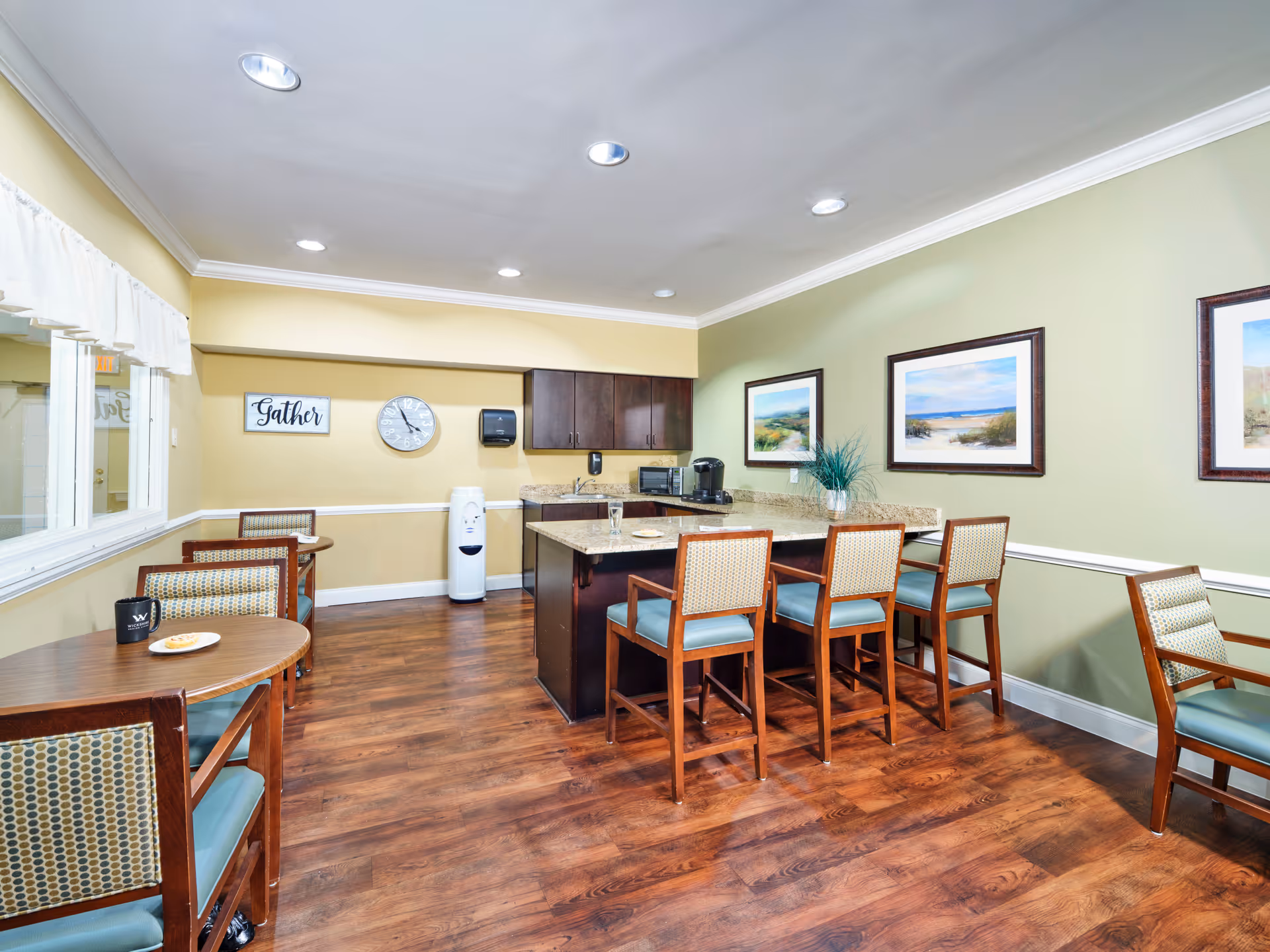 A bright and clean dining area with wooden floors, several wooden tables and chairs with patterned cushions, a kitchen island with four high chairs, and framed landscape paintings on the walls. The room has recessed lighting and a water dispenser in the corner. A sign on the wall reads 'Gather'.