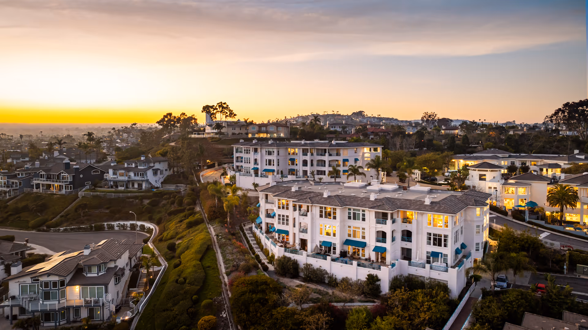 Aerial view of a residential community at sunset featuring multiple multi-story buildings with white exteriors and blue awnings, surrounded by greenery and other houses on a hillside.
