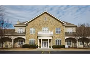 Front view of a two-story stone-faced senior living building with a central entrance, balcony, and surrounding landscaping.