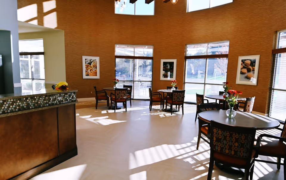 A bright dining area with round tables and chairs, each table decorated with a small vase of red flowers. Large windows with blinds allow natural light to fill the room. The walls are adorned with framed pictures of food items. A counter with a mosaic tile front is visible on the left side of the image.