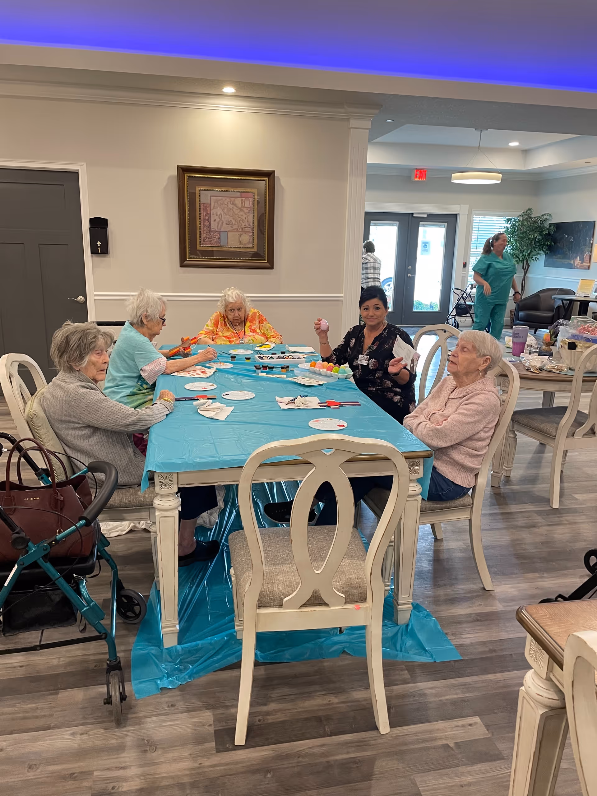 A group of elderly women sitting around a table covered with a blue plastic tablecloth, engaging in a craft activity involving colorful materials. The room has wooden flooring, light-colored walls, and framed artwork. In the background, a woman in green scrubs is walking near a seating area with chairs and a potted plant.
