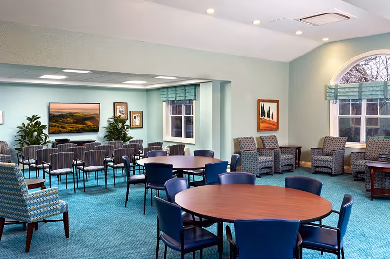 A spacious, well-lit common area with round wooden tables surrounded by blue chairs in the foreground and rows of striped chairs facing a wall-mounted TV in the background. The room has light green walls, large windows with green valances, framed artwork, and several upholstered armchairs arranged near the windows.
