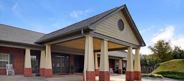 Exterior view of a senior living facility entrance with a covered drop-off area supported by columns, brick and beige siding, and a clear blue sky above.