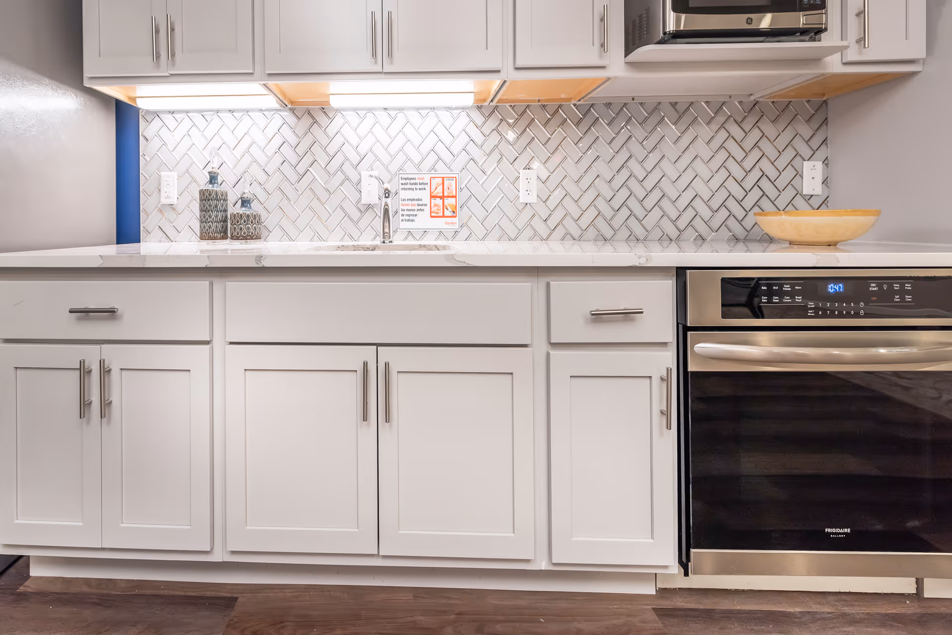 Modern kitchen countertop with white cabinets, a stainless steel oven, a microwave, a sink, decorative bottles, and a wooden bowl. The backsplash features a white herringbone tile pattern.