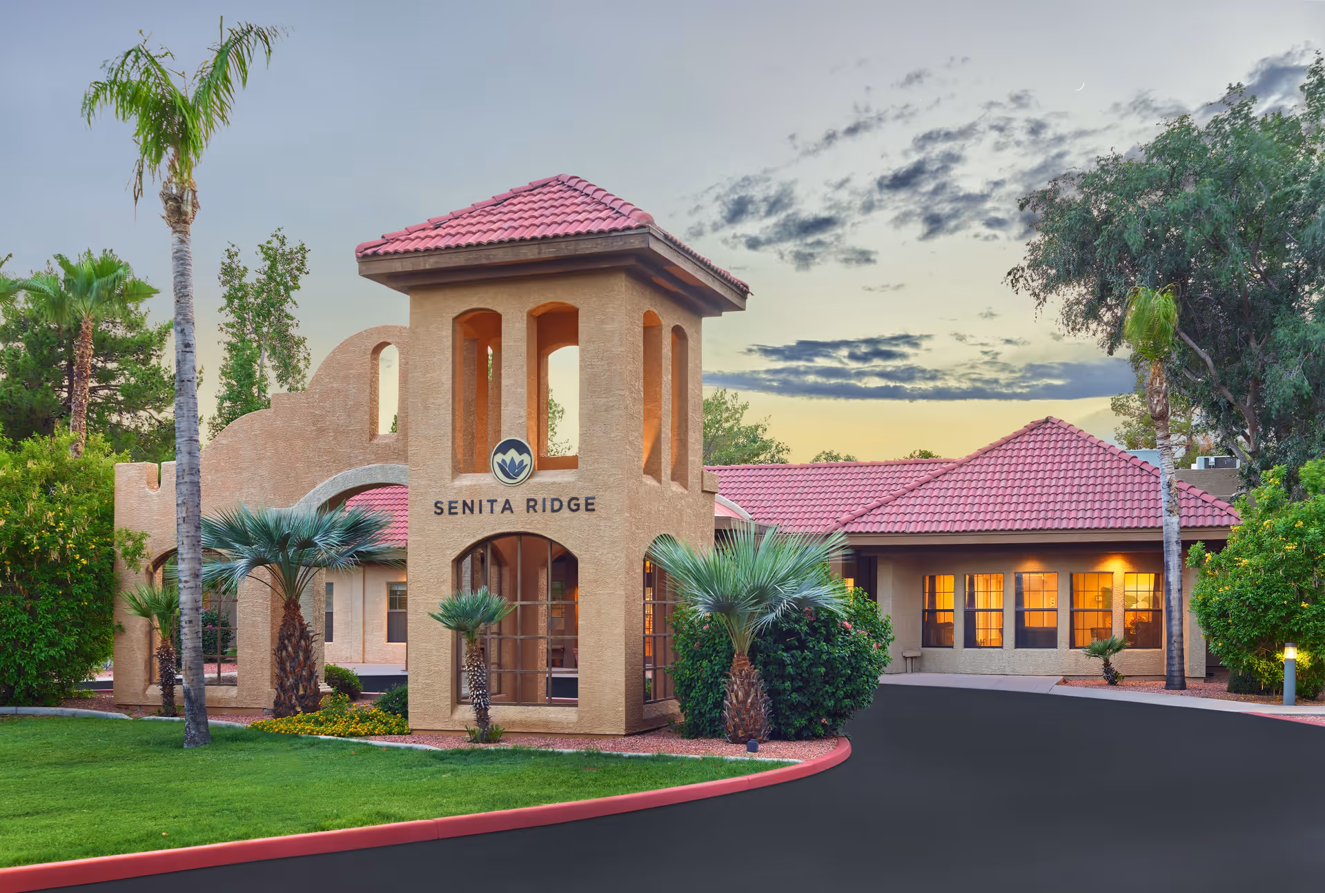 Exterior view of Senita Ridge facility at dusk, featuring a beige stucco building with a red tiled roof, arched windows, and a tower-like entrance structure. The surrounding landscape includes palm trees, green grass, and shrubs, with a paved driveway curving in front of the building.