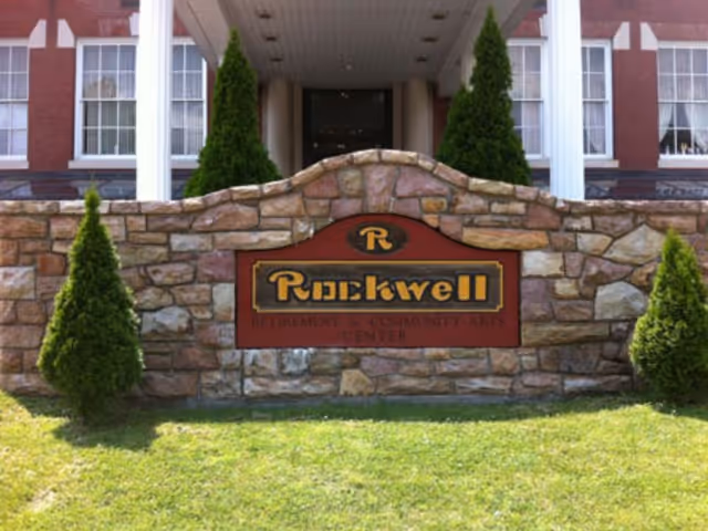 Stone entrance sign reading 'Rockwell' in front of a brick building with white columns and small evergreen shrubs.