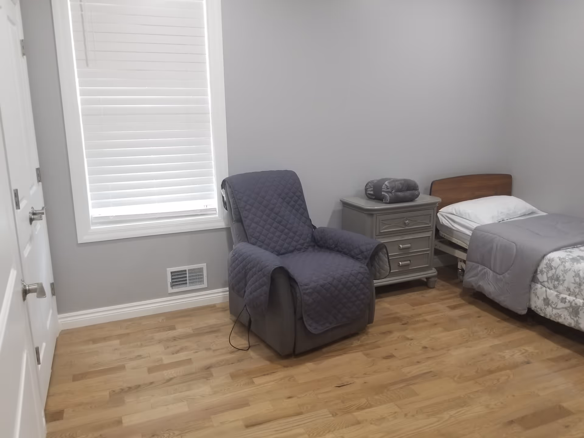 A simple bedroom with light gray walls and wooden floor. The room contains a single bed with a wooden headboard, white pillow, and gray bedding. Next to the bed is a gray nightstand with three drawers, on top of which are two folded gray blankets. A gray recliner chair with a quilted cover is positioned near a window with white blinds. There are two white doors on the left side of the room.