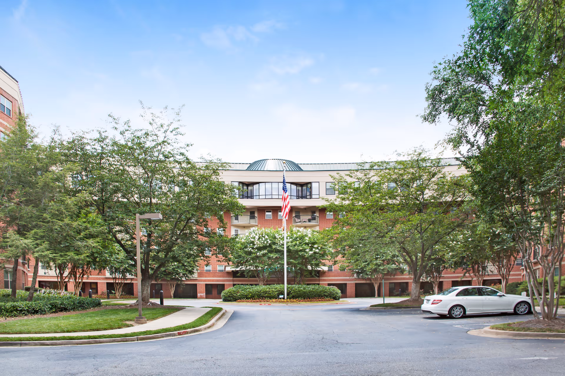 Front exterior of a multi-story brick senior living building with an American flag in the driveway, trees, and a parked car.
