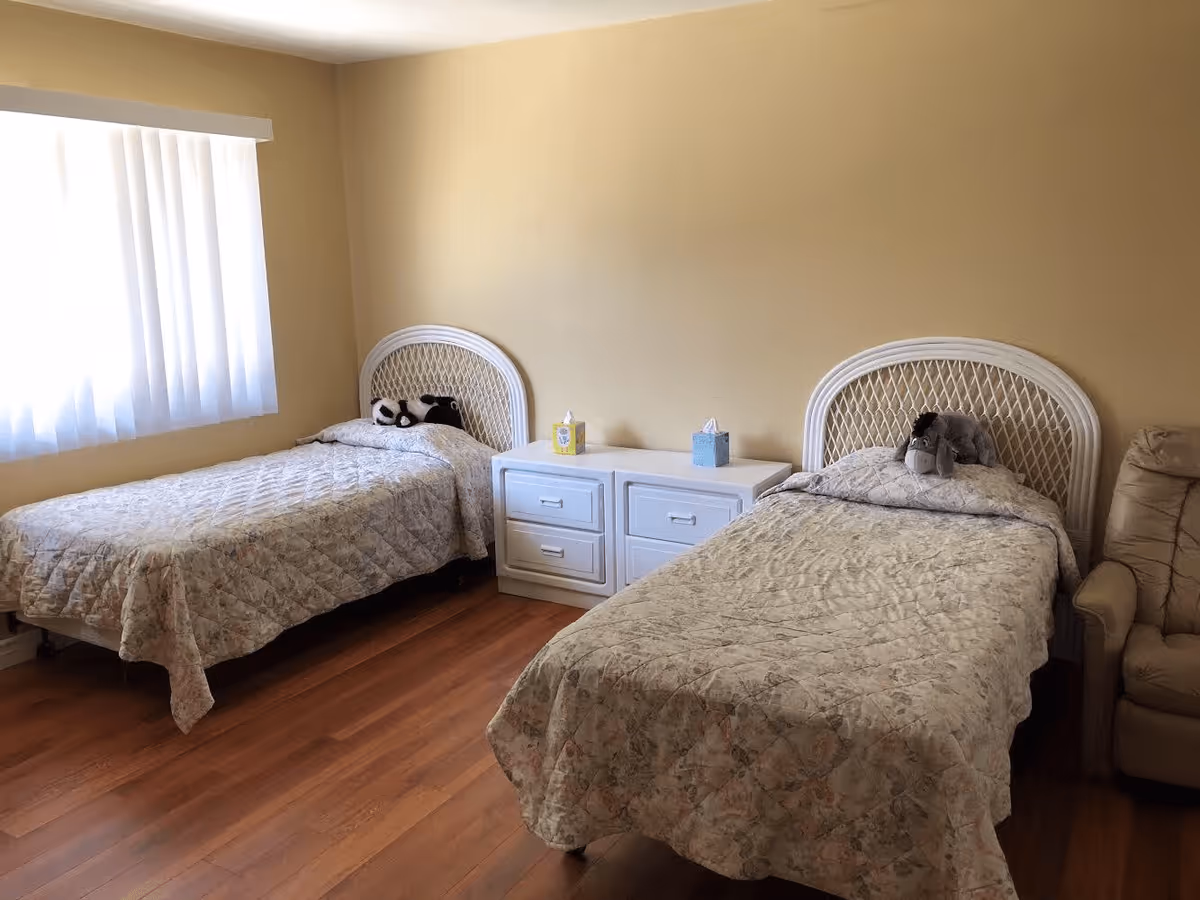 A softly lit bedroom with two twin beds featuring white wicker headboards, a shared nightstand between them, and a window with curtains.