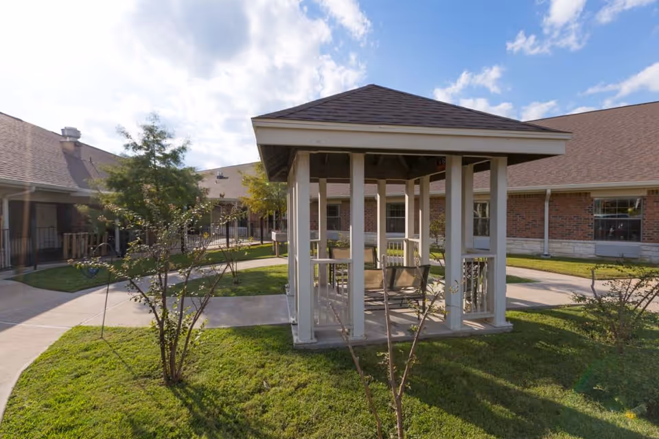 Outdoor courtyard area with a small gazebo structure featuring a bench inside, surrounded by grass, small trees, and a paved walkway. The background shows a brick building with multiple windows under a partly cloudy sky.