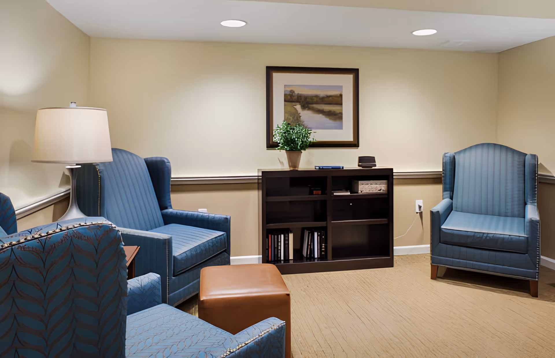 A cozy sitting room with blue upholstered armchairs, a lamp, ottoman, and a bookshelf topped with a framed landscape and potted plant.