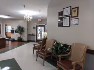 Interior view of a senior living facility hallway with three wooden chairs with cushioned seats and backs arranged against a wall. Above the chairs, several framed certificates and awards are displayed. There are potted plants near the chairs and in the hallway. The hallway leads to a glass door exit with an illuminated exit sign above it. The floor has a green and white pattern, and a chandelier hangs from the ceiling.
