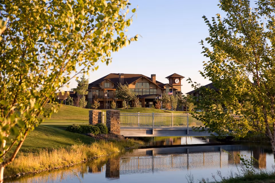 Exterior view of a senior living building with a clock tower, a bridge over a pond, and trees in the foreground.