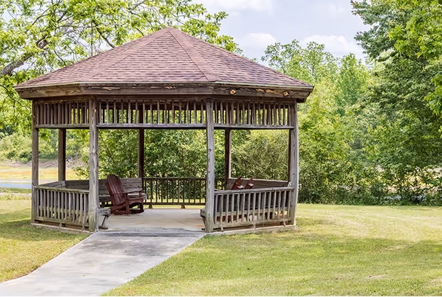 Wooden octagonal gazebo with chairs at the end of a concrete path in a grassy, tree-lined outdoor area.