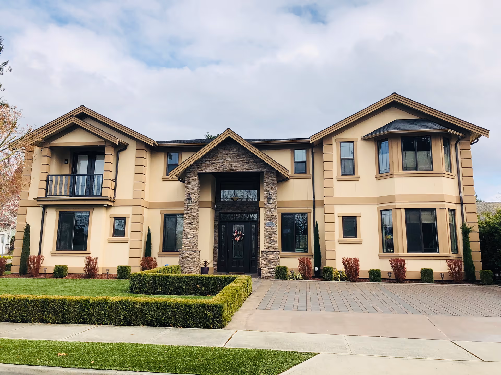 Front exterior view of a large two-story beige house with stone accents around the main entrance, multiple windows, a small balcony, and neatly trimmed hedges and lawn in front.