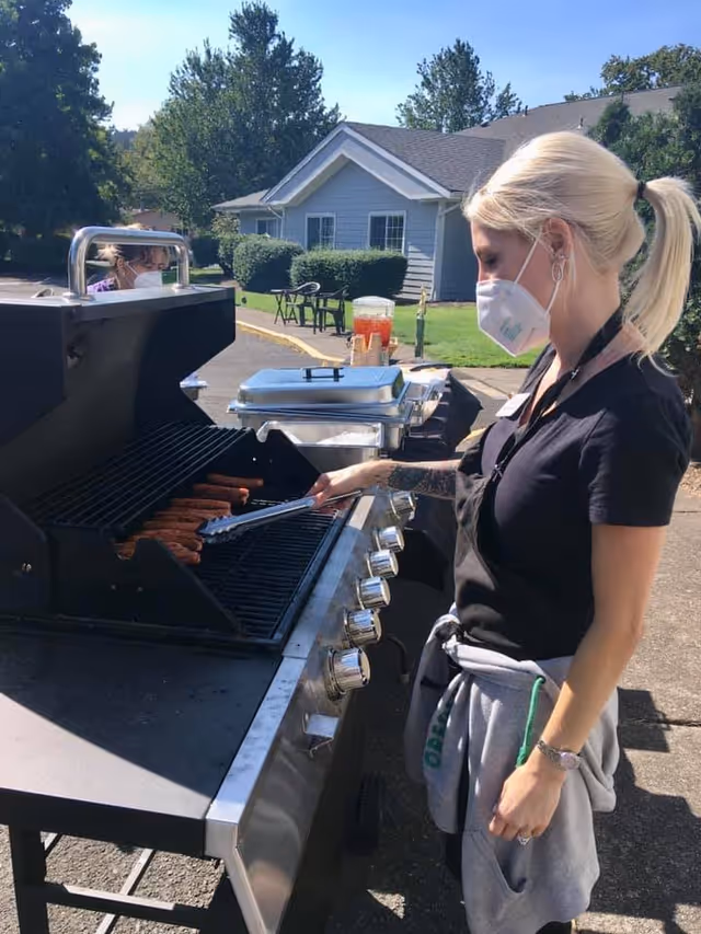 A woman wearing a face mask and black shirt is grilling sausages on a large outdoor gas grill. She is using tongs to turn the sausages. In the background, there is another person also wearing a mask, a table with covered trays and a beverage dispenser, and a residential-style building with bushes and trees under a clear blue sky.