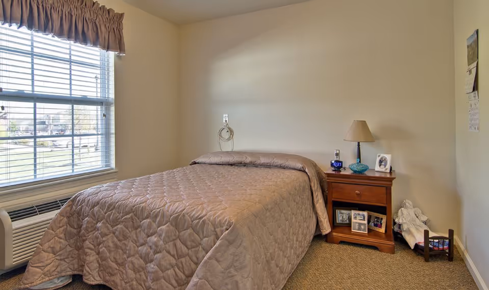 A simple bedroom with a bed covered in a beige quilted bedspread, a wooden nightstand with a lamp, alarm clock, and framed photos. There is a window with blinds and a valance on the left side, and a small figurine or statue on the floor near the nightstand.