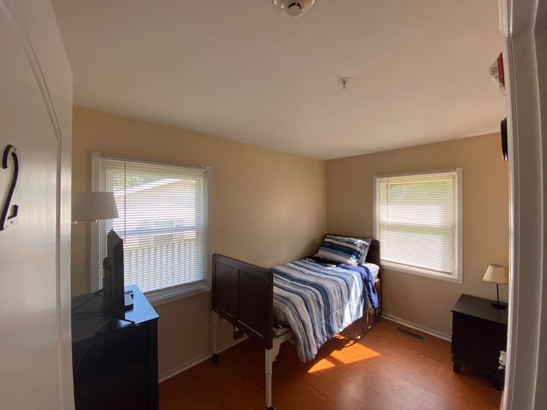A small, simple bedroom with beige walls and two windows with blinds. The room contains a single bed with a blue and white striped comforter and pillow, a black dresser with a TV on top, and a small black nightstand with a lamp. The door to the room is partially open and marked with the number 2.
