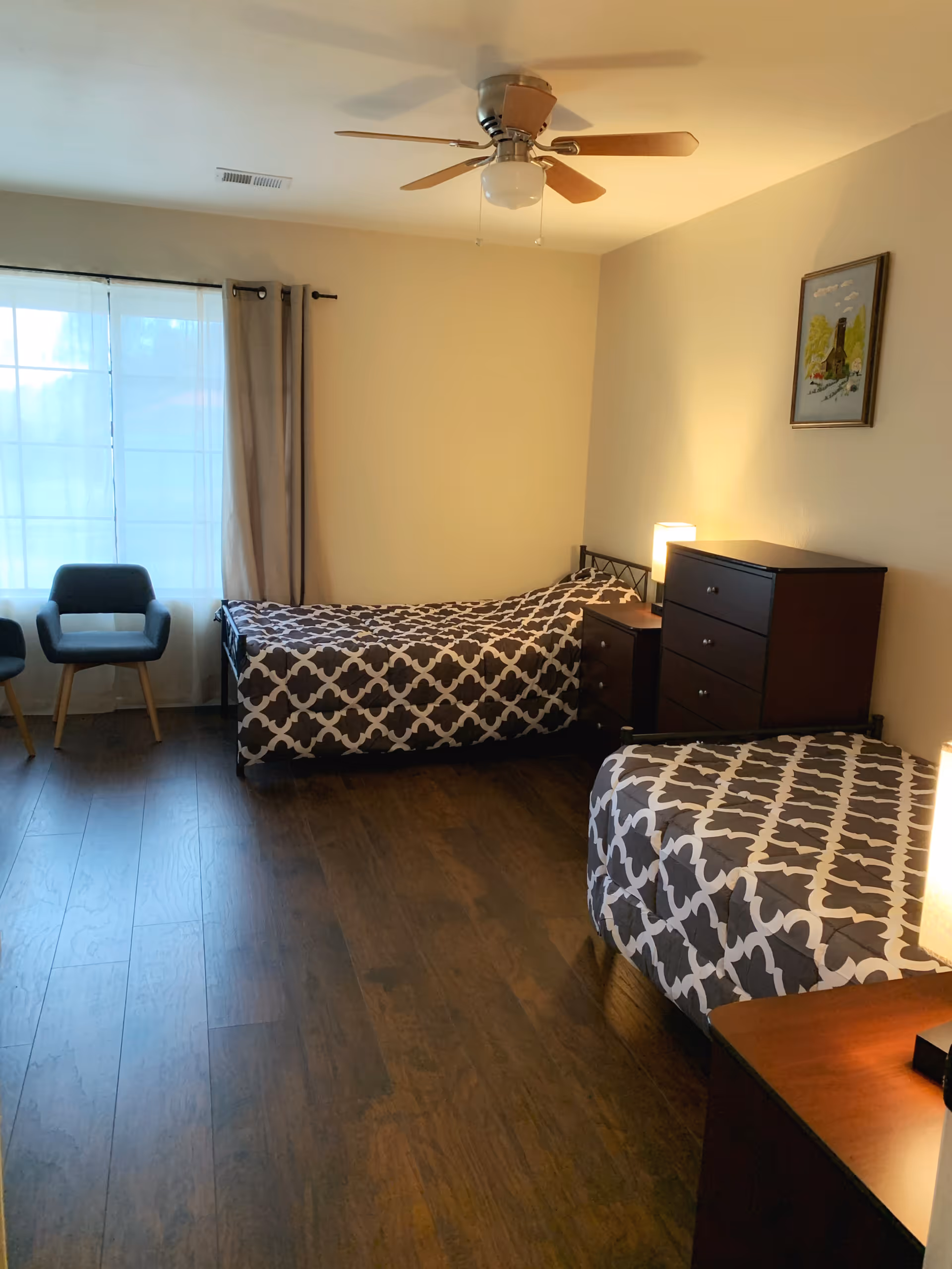 A bedroom with two single beds covered in matching patterned bedspreads. There are two dark wooden dressers between and beside the beds, each with a lamp on top. A ceiling fan with wooden blades is mounted on the ceiling. Two gray chairs are positioned near a window with sheer white curtains and beige drapes. A framed picture hangs on the wall above one of the dressers. The floor is dark wood.