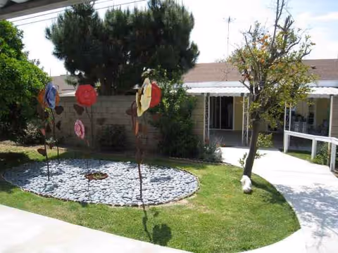 Outdoor courtyard with a circular rock garden, trees, and a walkway beside a single-story building.