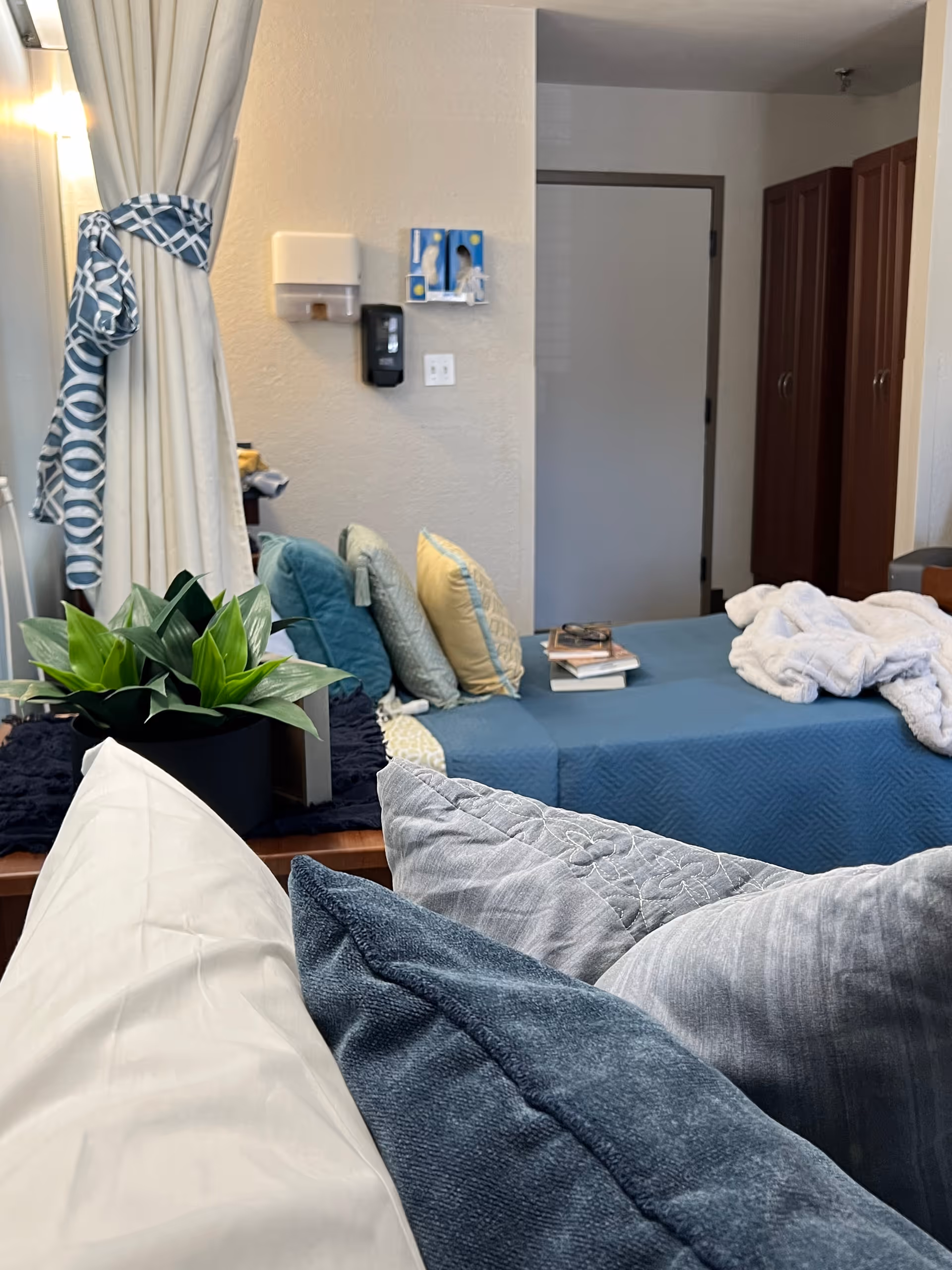 A cozy bedroom in a senior living facility with a bed covered in blue bedding and multiple pillows in shades of blue and gray. There is a small plant on a wooden surface near the bed, a curtain tied back with a patterned tie, and a white door in the background. Some books and a white blanket are on the bed, and there are cabinets along the wall.