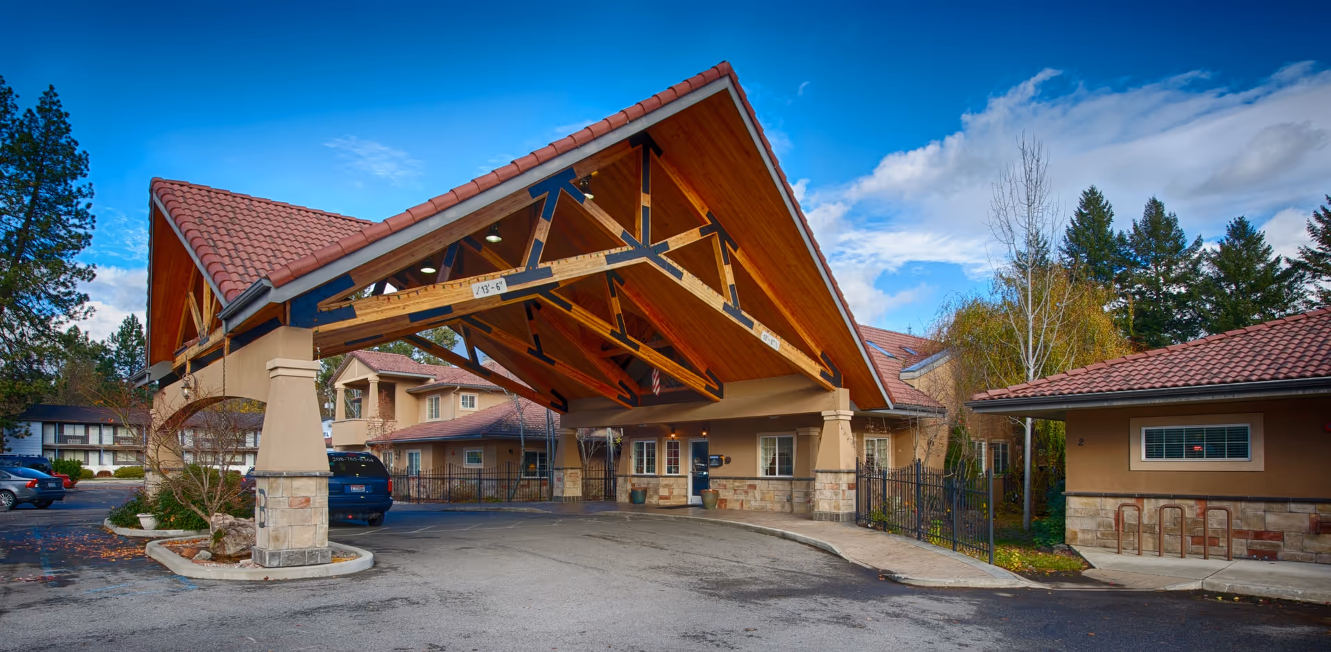 Entrance of a senior living facility with a large wooden canopy supported by stone pillars, surrounded by buildings with red-tiled roofs and trees under a partly cloudy blue sky.