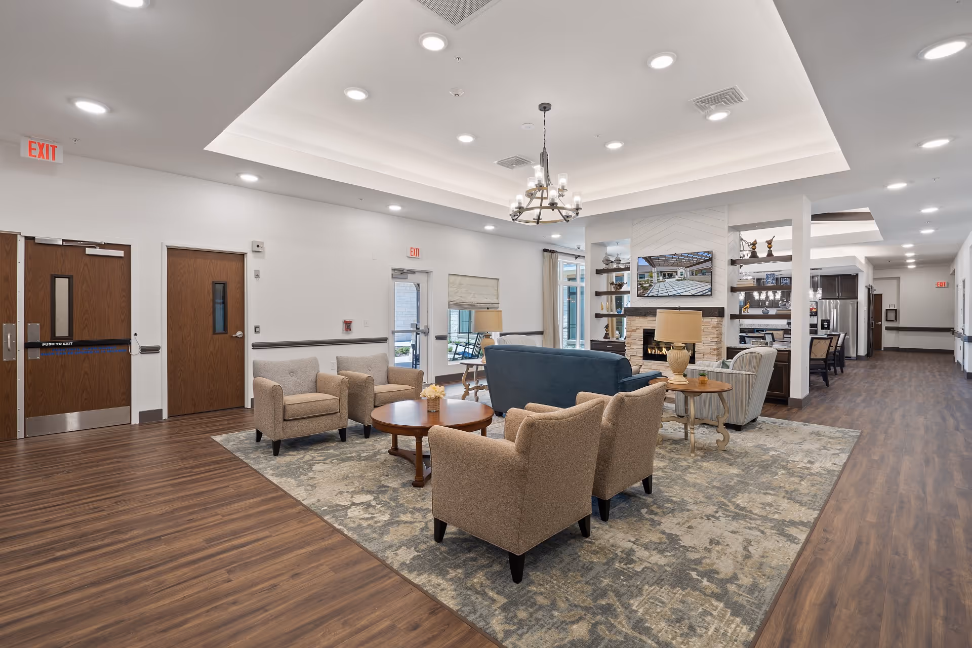 A spacious and well-lit senior living common area with comfortable seating including beige armchairs and a blue sofa arranged around a wooden coffee table on a patterned rug. The room features a modern fireplace with a mounted TV above it, decorative shelves, and a chandelier hanging from a recessed ceiling. The flooring is wood, and there are multiple exit doors and windows letting in natural light.