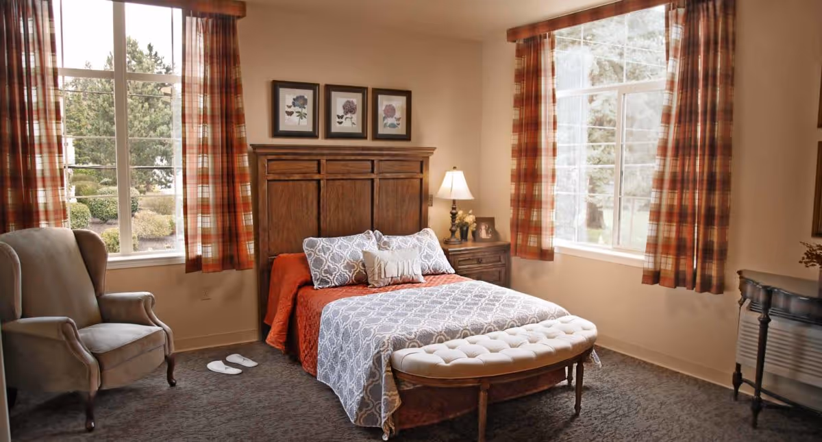 A cozy bedroom with a wooden headboard bed dressed in red and gray patterned bedding, two large windows with plaid curtains letting in natural light, a cushioned bench at the foot of the bed, a beige armchair to the left, and a wooden nightstand with a lamp and framed photo on the right.