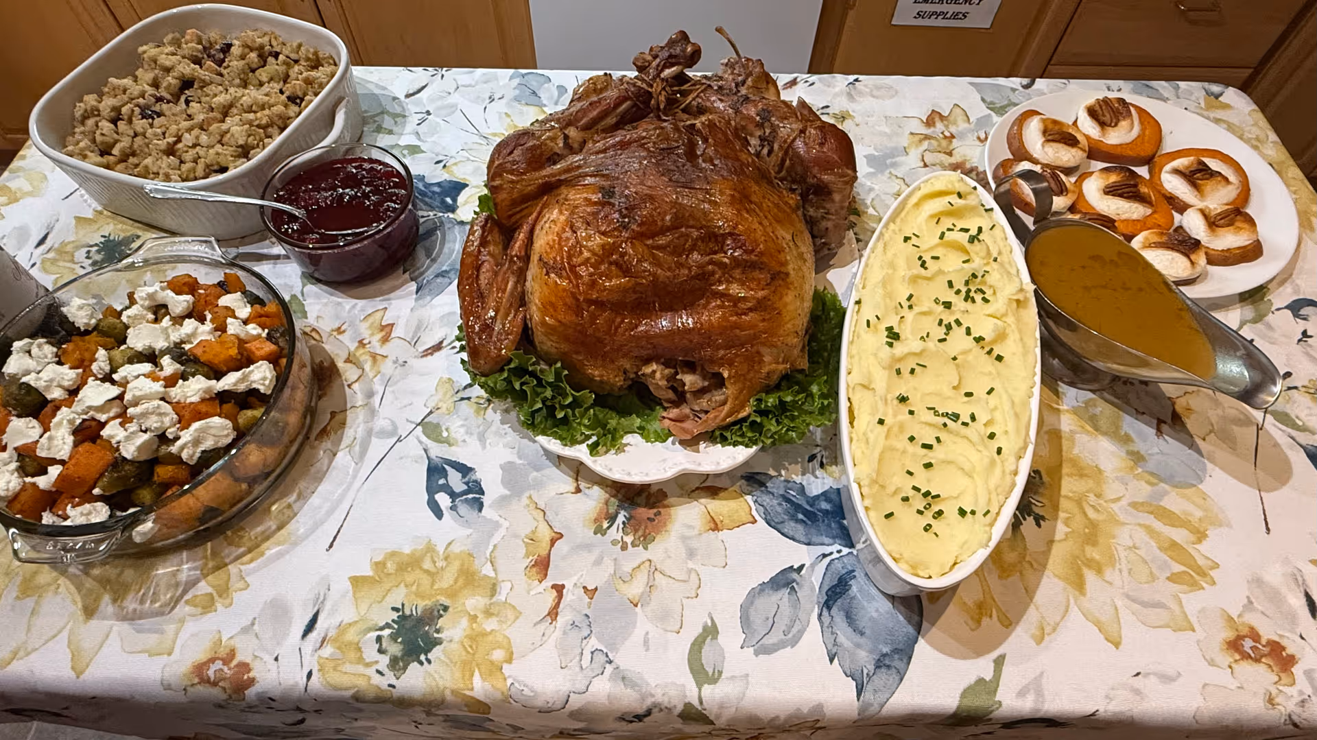 A table covered with a floral tablecloth displaying a roasted whole turkey on a bed of lettuce, a dish of mashed potatoes garnished with chopped chives, a gravy boat filled with brown gravy, a bowl of cranberry sauce, a casserole dish with a crumble topping, a glass dish with roasted vegetables topped with dollops of white cheese, and a plate of small pumpkin-shaped desserts with toasted marshmallows on top.