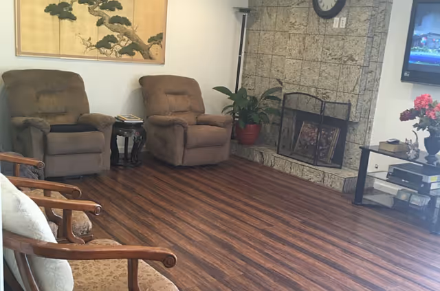 A cozy living room with two brown recliner chairs, a wooden armchair with a cushion, a stone fireplace with a black metal screen, a potted plant in a red pot, a wall clock above the fireplace, a flat-screen TV mounted on the wall, and a glass TV stand with decorative items and flowers.