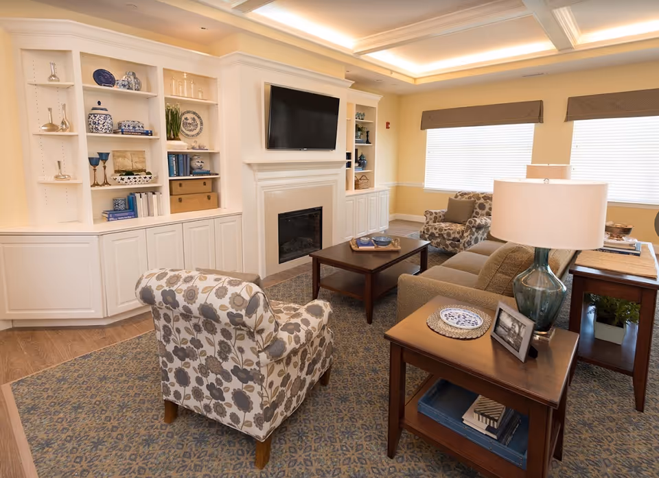 Bright furnished living room with patterned armchairs, a sofa, coffee table, wall-mounted TV above a fireplace, and built-in shelving.