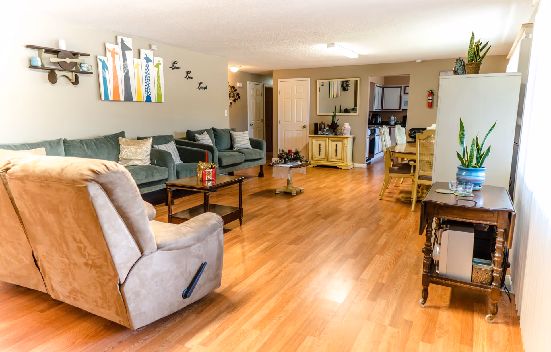A cozy living room with wooden flooring, featuring a beige recliner chair in the foreground, two green sofas with cushions along the left wall, a wooden coffee table with a decorative gift box, and a small round table with a floral centerpiece. The wall has colorful giraffe artwork and the words 'Love Live Laugh'. In the background, there is a yellow cabinet with plants and a mirror above it, leading to a dining area with a wooden dining table and chairs, and a kitchen visible beyond.