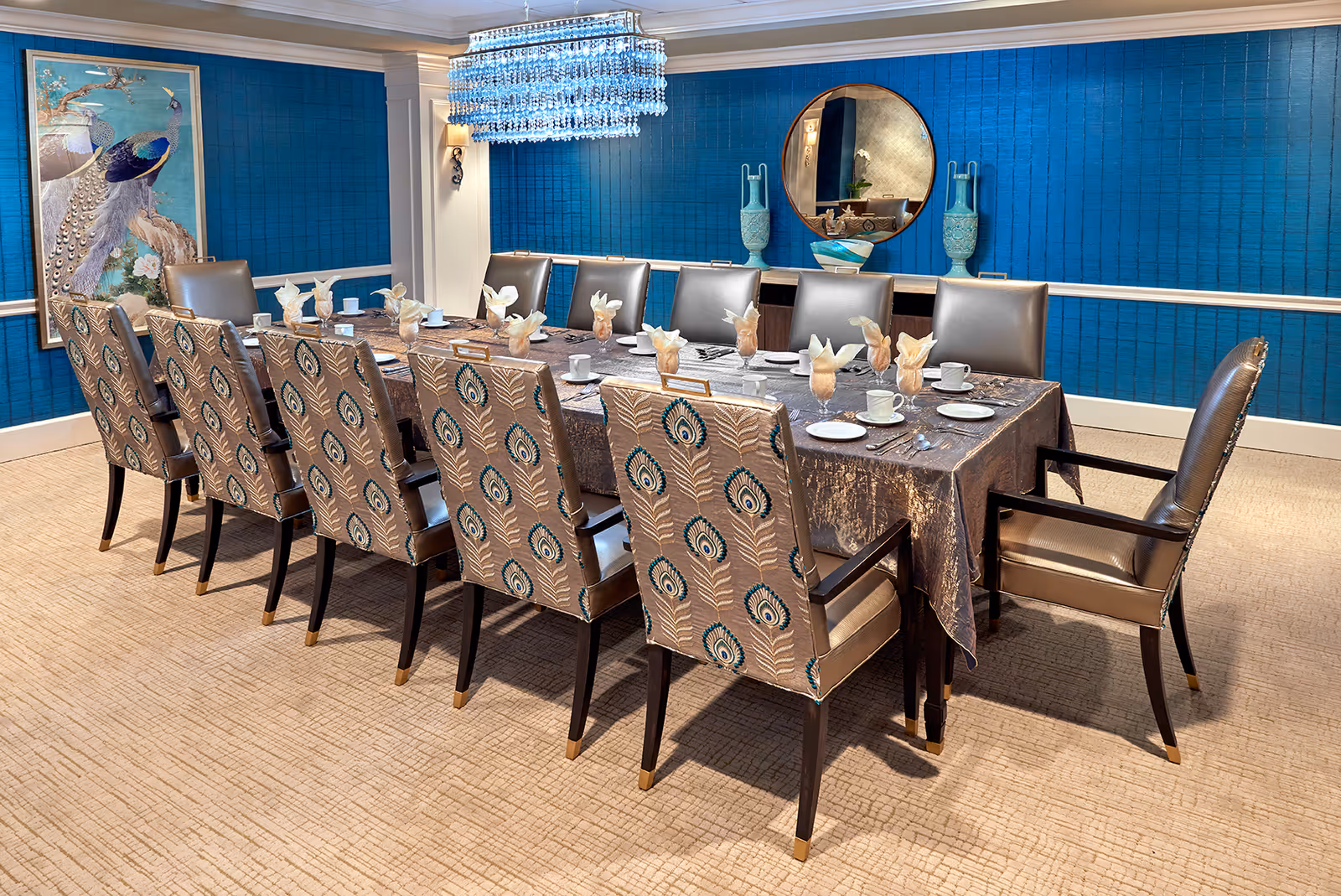 A formal dining room with a long rectangular table covered with a dark tablecloth, set with cups, saucers, plates, and napkins folded in a decorative manner. The room features twelve upholstered chairs with peacock feather patterns on the backrests and two leather armchairs at the ends of the table. The walls are painted blue with white trim, and there is a large framed peacock artwork on one wall and a round mirror with decorative vases on a sideboard. A modern crystal chandelier hangs above the table.